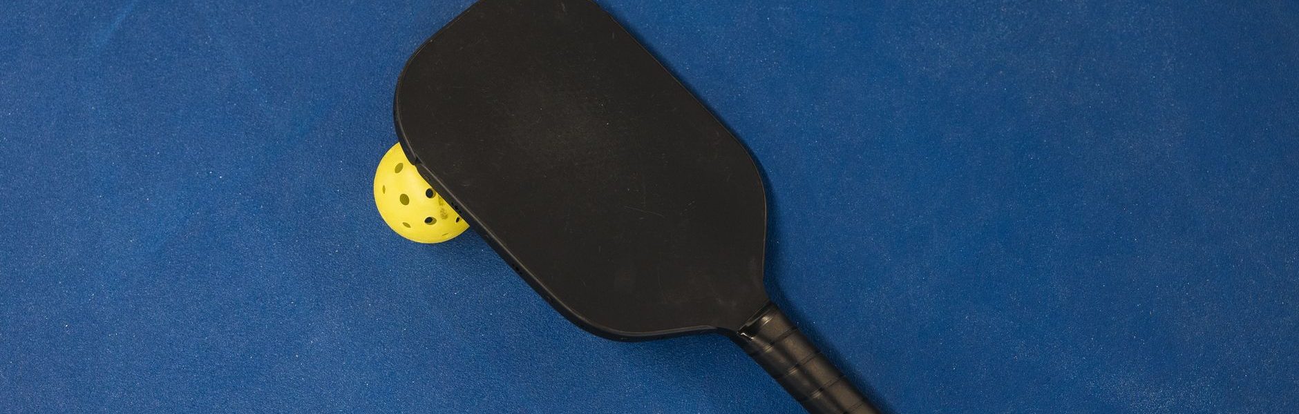 Black pickleball paddle resting on a yellow pickleball on a blue surface