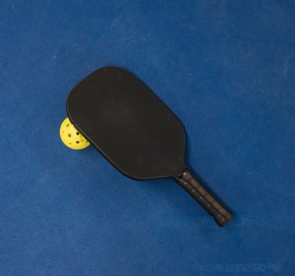 Black pickleball paddle resting on a yellow pickleball on a blue surface